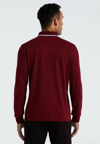 Man die met zijn rug naar de kijker staat, draagt een donkerrode polo met lange mouwen en een witte streep op de kraag, in combinatie met zwarte broek.