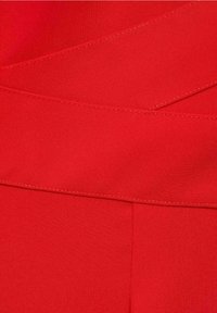 Un tissu rouge avec une texture lisse présente un design superposé et des coutures visibles. Le produit met en avant une taille ajustée et des plis plats.