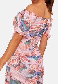 Nainen pukeutuneena pastellinsävyiseen kukalliseen olkapäät paljastavaan mekkoon, jossa on pussihihaiset ja rypytetyt yksityiskohdat. Näkyvissä on hänen selkänsä, osa käsivartta ja hiuksia.