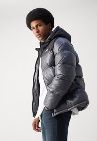 Dunjakke i mørkegrått, med hette, glidelåslommer og en blank overflate. Kombinert med en hvit skjorte og blå jeans.