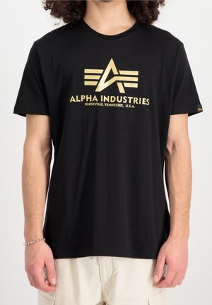 Czarna koszulka z złotym logo Alpha Industries i napisem "Knoxville, Tennessee, U.S.A.", noszona przez osobę o jasnej karnacji i kręconych włosach.