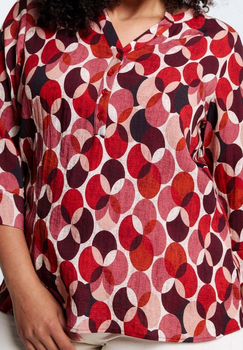Donna che indossa una blusa a motivi rossi, rosa e neri con forme circolari e colletto a V con bottoni.