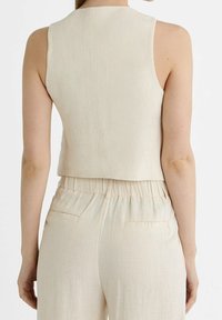 Korte mouwloze top in lichtbeige, gemaakt van textuurstof. Heeft een hoge halslijn en een gladde achterkant. Gecombineerd met een broek met elastische tailleband.