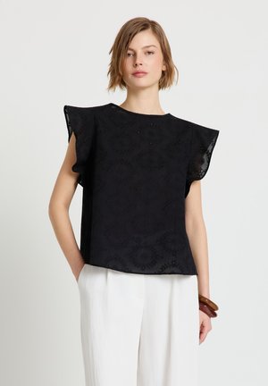 Giovane donna che indossa una blusa nera senza maniche con spalle arruffate e pantaloni bianchi a gamba larga, in piedi con una mano in tasca.