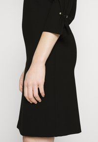 Vue latérale d'une personne portant une robe noire longueur genou avec des manches trois-quarts présentant des détails de boutons près du coude.