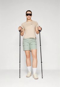 Bēša īsroku krekls, gaiši zaļas kravas šorti un pārgājienu nūjas. Modelis valkā tumšas saulesbrilles un gaišas kurpes ar baltām zeķēm.