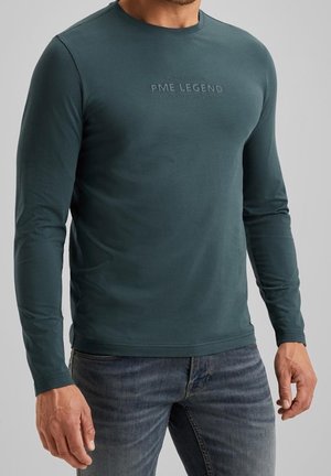 Homme portant une chemise manches longues PME Legend vert foncé avec logo embossé et jean bleu délavé sur fond gris uni.
