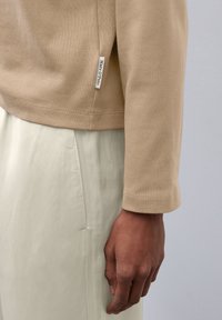 Beige Baumwolloberteil mit langen Ärmeln und seitlichem Schlitz, kombiniert mit hellgrünen Hosen. Verfügt über ein kleines Markenetikett am Saum.