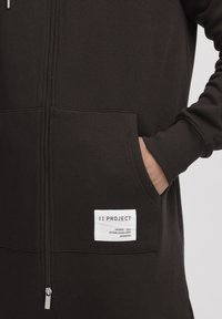 Black zip-up hoodie made of soft cotton blend. Features side pockets, ribbed cuffs, and a white logo label on the front.