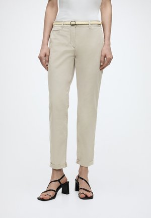 Frau trägt beige hochgekrempelte Hose mit geflochtenem Gürtel und schwarze Riemchen-Sandalen mit hohem Absatz, stehend vor weißem Hintergrund.