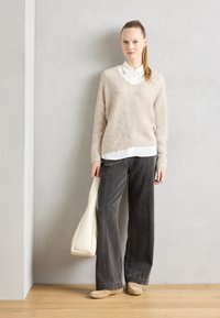 Beige strikkegenser, hvit skjorte under, grå vide bukser, lyse flate sko, holder en hvit håndveske, minimalistisk bakgrunn.