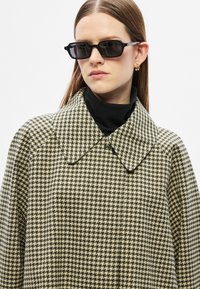 Manteau à motif pied-de-poule en vert et crème, avec un grand col et une fermeture à boutons. Porté sur un col roulé noir. Des lunettes de soleil noires complètent le look.