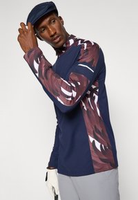 Long-sleeve athletic top in navy with a patterned maroon and white design on sleeves, featuring a half-zip collar and modern fit.