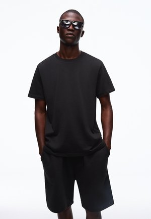 Mężczyzna w czarnych okularach przeciwsłonecznych, czarnej koszulce z krótkim rękawem i czarnych szortach, stoi z rękami w kieszeniach na białym tle.