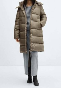 Doudoune vert olive, tissu brillant, longueur mi-cuisse, fermeture éclair et boutons-pression, large capuche, portée sur une robe grise fendue.