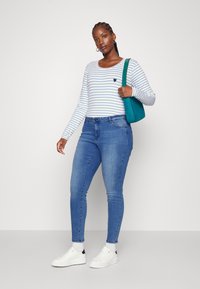 Long-sleeve striped shirt with a small heart emblem, paired with blue skinny jeans and white sneakers, accompanied by a teal handbag.