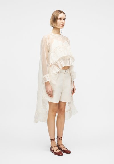 Γυναίκα που φοράει διαφανές λευκό τοπ με βολάν και μακριά μανίκια, λευκά σορτς και καφέ επίπεδα παπούτσια με κορδόνια, στέκεται μπροστά σε απλό λευκό φόντο.