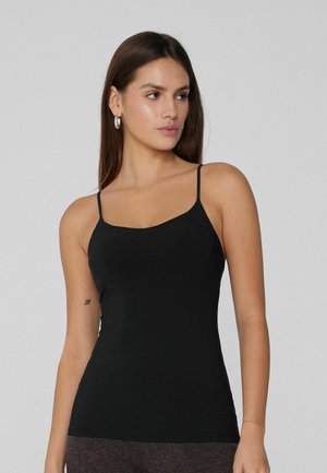 Donna con lunghi capelli scuri indossa un top nero con spalline sottili, orecchini a cerchio d'argento e pantaloni grigi, in piedi davanti a uno sfondo chiaro e semplice.