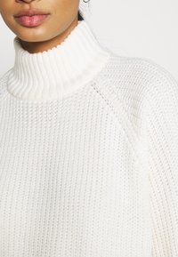 Maglione spesso bianco a maglia con colletto alto a coste e maniche raglan, che presenta un motivo testurizzato e un tessuto morbido.