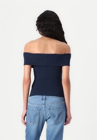 Jūrinės spalvos off-shoulder palaidinė su prigludusia forma, pagaminta iš lygaus audinio. Suderinta su šviesiai mėlynomis džinsinėmis kelnėmis, turinčiomis užpakalines kišenes ir šiek tiek išblukimu.