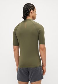 Rash guard s krátkym rukávom v olivovo zelenej farbe, so bezšvovým dizajnom a logom v oranžovej farbe na chrbte. Vyrobené z pružného, priedušného materiálu.