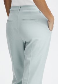 Primer plano de una persona que lleva pantalones azul claro a medida, con la mano en el bolsillo lateral, mostrando detalles del bolsillo trasero y lateral.