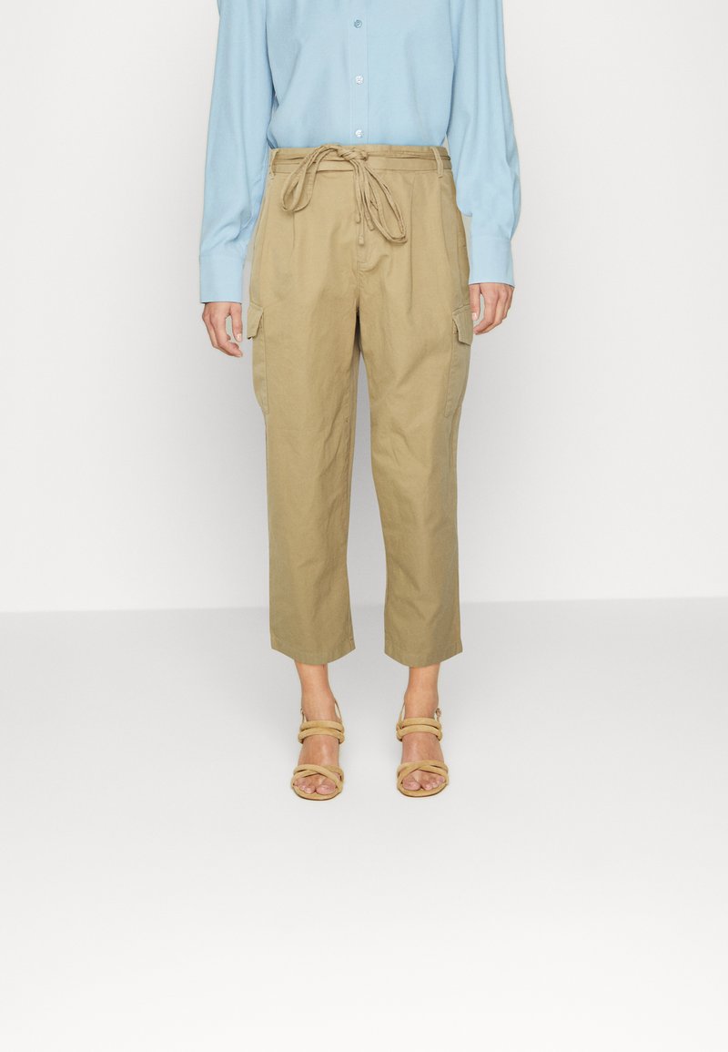 Calças cargo caqui com cintura ajustável, comprimento cropped e bolsos laterais, combinadas com uma camisa de mangas compridas azul claro e sandálias bege.