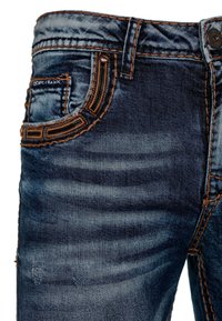 Jean en denim avec un délavage bleu foncé, présentant des coutures orange contrastantes, des poches avant et des détails usés sur le tissu.