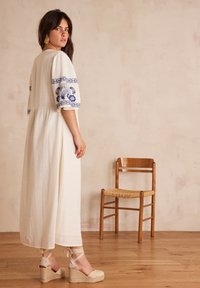 Abito lungo bianco con maniche ricamate in blu, in tessuto leggero e silhouette fluida. Sandali wedge con texture intrecciata su un pavimento in legno.