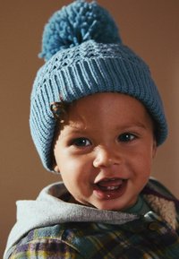 Bonnet en tricot bleu avec un pompon sur le dessus, présentant un motif texturé. Un enfant porte une veste à carreaux avec une capuche et une chemise de couleur claire en dessous.