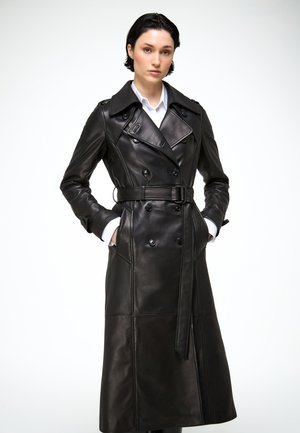 Vrouw die een lange zwarte leren trenchcoat met riem en wit shirt draagt, staand met handen in de zakken tegen een witte achtergrond.