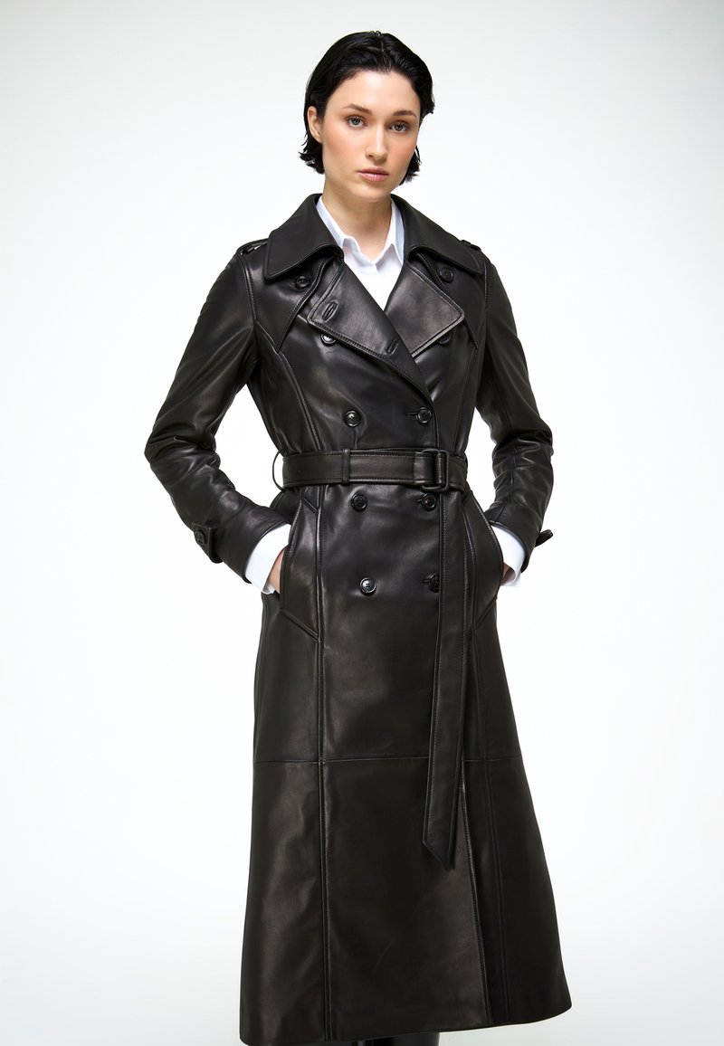 Femme portant un long trench-coat en cuir noir avec ceinture et une chemise blanche, debout les mains dans les poches devant un fond blanc.
