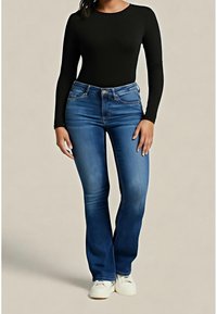 Femme vêtue d'un haut noir ajusté à manches longues, d'un jean bootcut bleu et de baskets blanches, debout devant un fond uni.