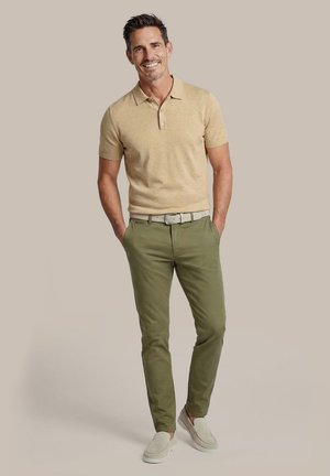 Man staat met handen in de zakken, draagt een beige polo met korte mouwen, olijfgroene broek, lichte riem en beige instapschoenen, glimlachend.