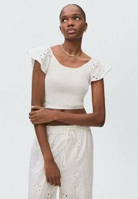 Top bianco corto con maniche ruffle corte e dettagli in pizzo. Abbinato a pantaloni bianchi che presentano motivi ritagliati floreali e cordoncino.