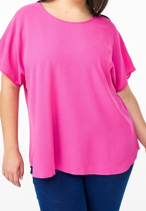 Femme portant un haut ample manches courtes rose vif et un pantalon bleu foncé, vue du cou aux hanches sur fond blanc.
