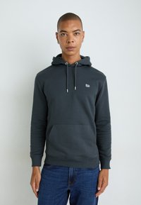 Donkergrijze hoodie met een voorzak, trekkoordcapuchon en een klein wit logo. Het materiaal lijkt zacht en textuurrijk; gecombineerd met blauwe jeans.
