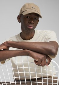 Căciulă bej cu un logo, tricou bej deschis, piele texturată, brațe odihnindu-se pe un scaun cu modele în grilă albă, fundal neutru.