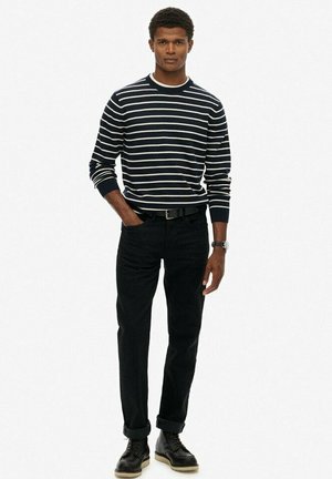 Mann in einem schwarz-weiß gestreiften Langarmshirt, schwarzen hochgekrempelten Hosen, schwarzem Gürtel, schwarzen Stiefeln und einer Armbanduhr, der vor einem weißen Hintergrund steht.