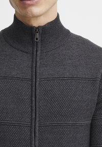 Gilet zippé gris foncé avec col à côtes et panneaux texturés. Il présente une fermeture éclair lisse et un tissu en tricot avec un détail de motif tissé.