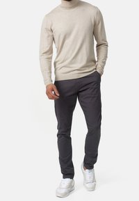 Beige Rollkragenpullover kombiniert mit dunkelgrauen Slim-Fit-Hosen und weißen Sneakers. Der Pullover hat eine glatte Textur und enganliegende Ärmel.