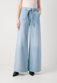 Světle modré kalhoty s širokými nohavicemi a zavazovacími vazy, s předními kapsami a uvolněným střihem. Materiál je denim s hladkou texturou.