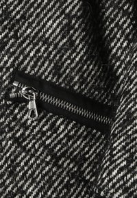 Tejido de pata de gallo en blanco y negro con una superficie texturizada, que presenta un bolsillo con cremallera negra en la esquina inferior izquierda.
