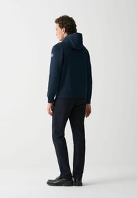 Hanorac bleumarin cu siret, manșete și tiv curbate, purtat cu pantaloni bleumarin și pantofi negri, văzut din spate.