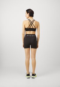 Reggiseno sportivo nero con spalline incrociate, abbinato a pantaloni corti neri a vita alta con logo bianco del marchio. Il modello indossa sneaker nere e verdi.
