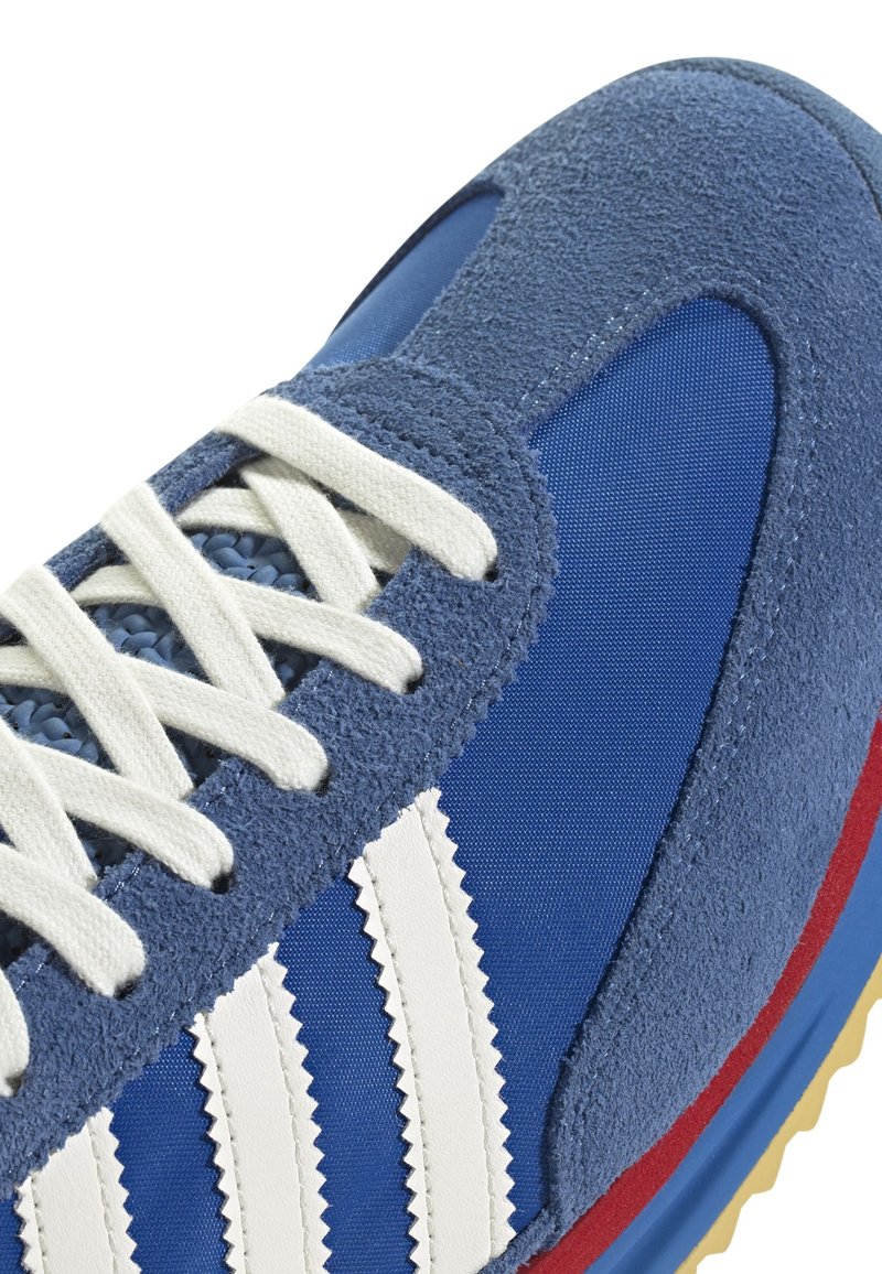 Basket bleue en daim et textile, avec des rayures blanches, un accent rouge et des lacets texturés. Bout arrondi et semelle en caoutchouc visibles.