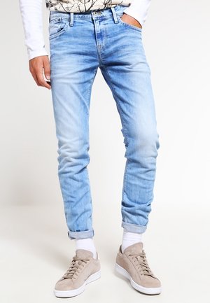 Lichtblauwe slim-fit jeans met omgeslagen boorden, gecombineerd met witte sokken en beige sneakers, gedragen door een persoon met één hand in de zak.