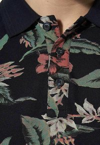 Bloemenpoloshirt met een zwarte achtergrond, voorzien van kleurrijke plantenpatronen, een gestructureerde stof en drie zwarte knopen bij de kraag.