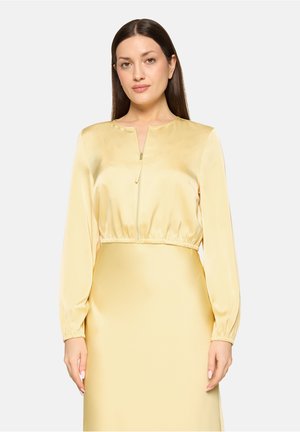 Frau mit langen dunklen Haaren, die ein hellgelbes, satinartiges Zip-up-Oberteil mit langen Ärmeln und einen passenden Rock trägt, steht vor einem schlichten Hintergrund.