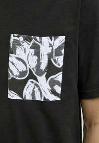 Camiseta negra con un bolsillo blanco en contraste que presenta un patrón de hojas en blanco y negro, mangas cortas y un corte clásico.